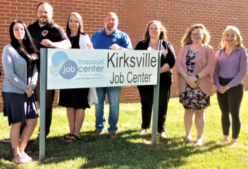 A group of people standing behind the Kirksville Job Center sign. They are all looking towards the camera and the sun is shining at them from behind the people on the right. 