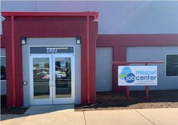The front of the Rolla Job Center. There is a door and sidewalk with a large rectangular red facade outlining the door and some front panels of the building. There is a Missouri Job Center sign hanging from some poles of the same red as the building.